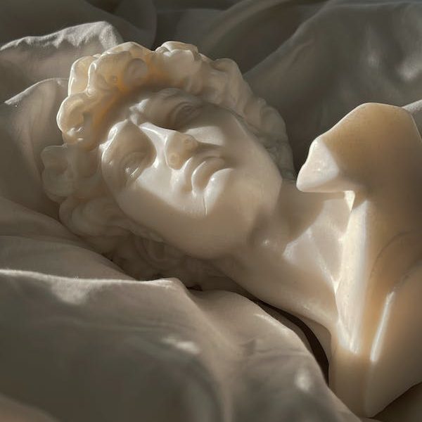 a white-marble-like head bust lying on white bedsheets while sunlight dapples the scene.